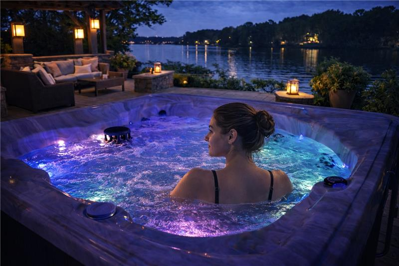 A woman soaking in a spa at dusk beside a lake, with the water glowing purple and teal from the LED lighting